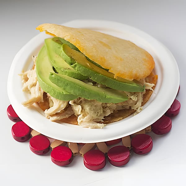 A Venezuelan La Arepa with shredded chicken and sliced avocado on a white plate.