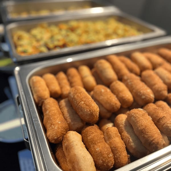 Golden-brown croquettes in a metal serving tray, with another tray of food blurred in the background.