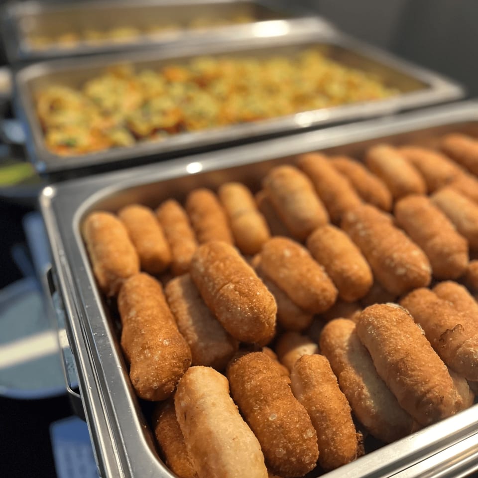 Golden-brown croquettes in a metal serving tray, with another tray of food blurred in the background.