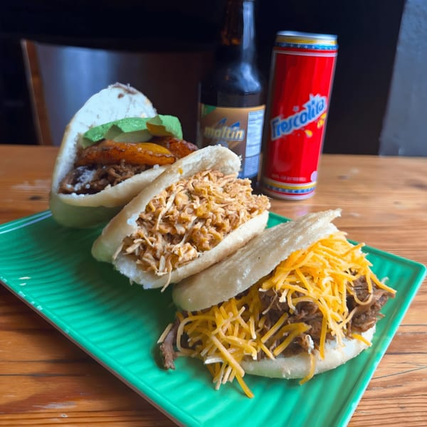 Three Venezuelan arepas with various fillings, a Maltin bottle, and a Frescolita can on a green plate.