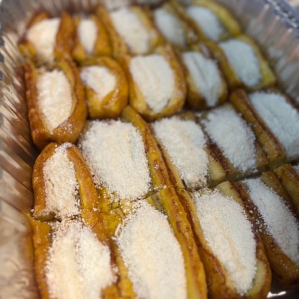 Rows of golden-brown plantains, sliced lengthwise and topped with white shredded cheese, in a foil tray.